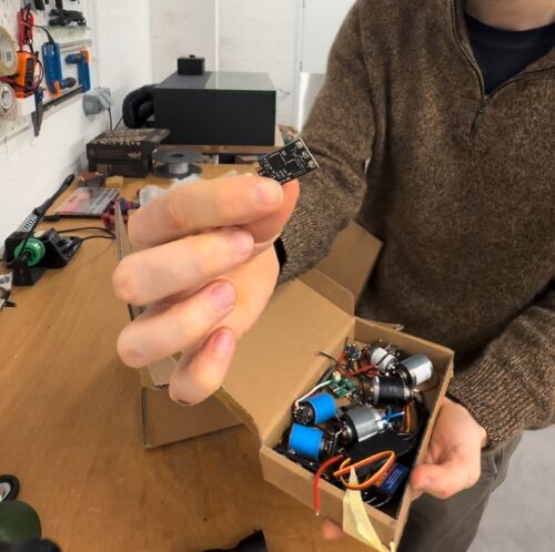 A hand holding up a small circuit board above an open box of assorted BLDC motors and electronic components on a workshop bench.