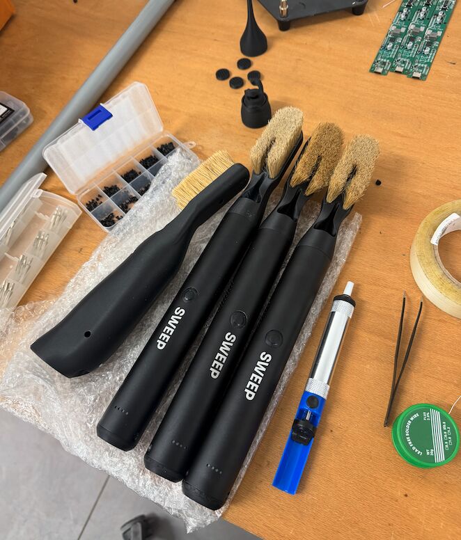 The first three ChalkBlaster units with SWEEP logos on a workbench, surrounded by brush heads, screws, a soldering iron and circuit boards