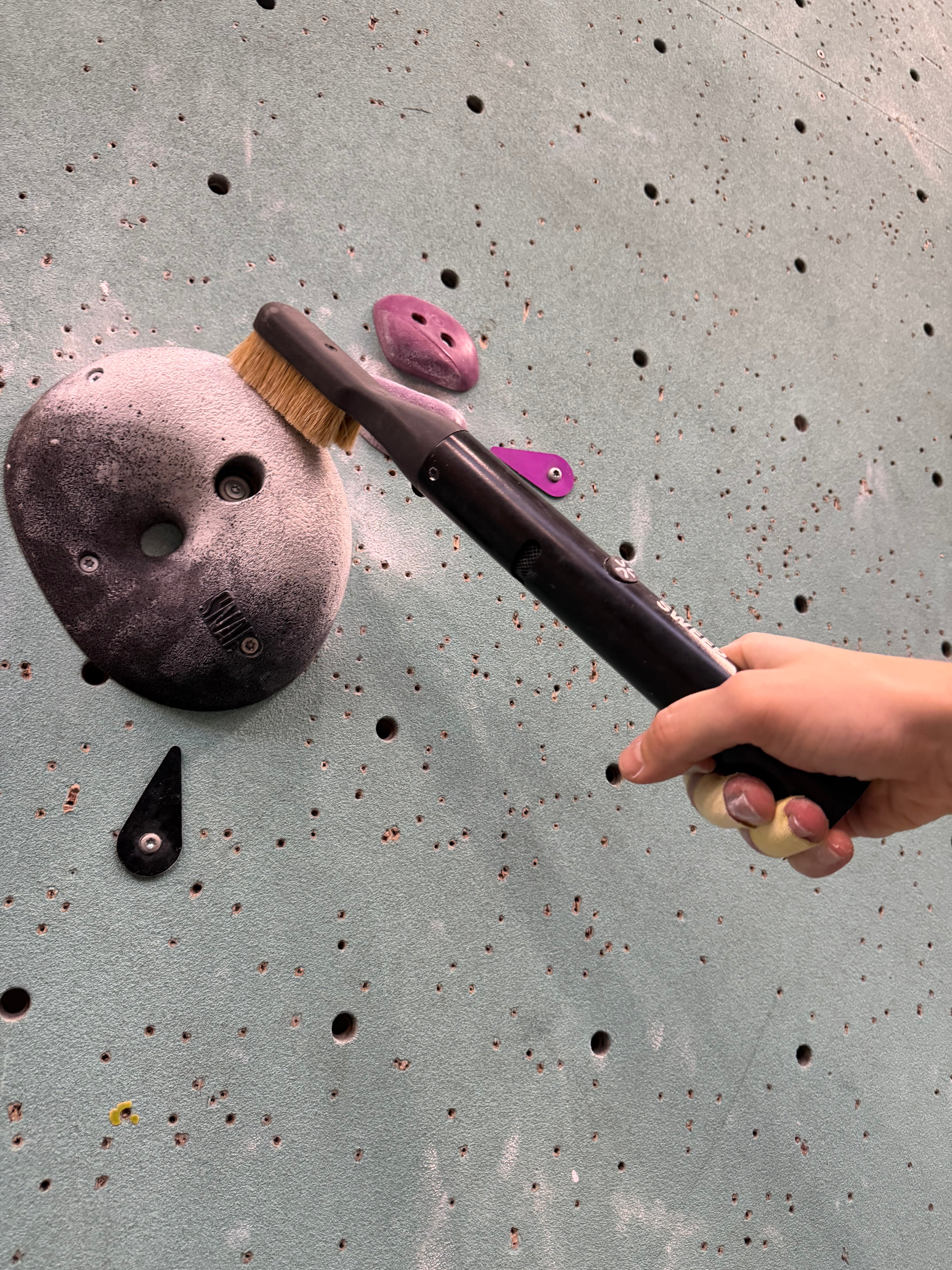 A bouldering climber holding a climbing brush in hand over a chalky climbing hold on a textured wall.